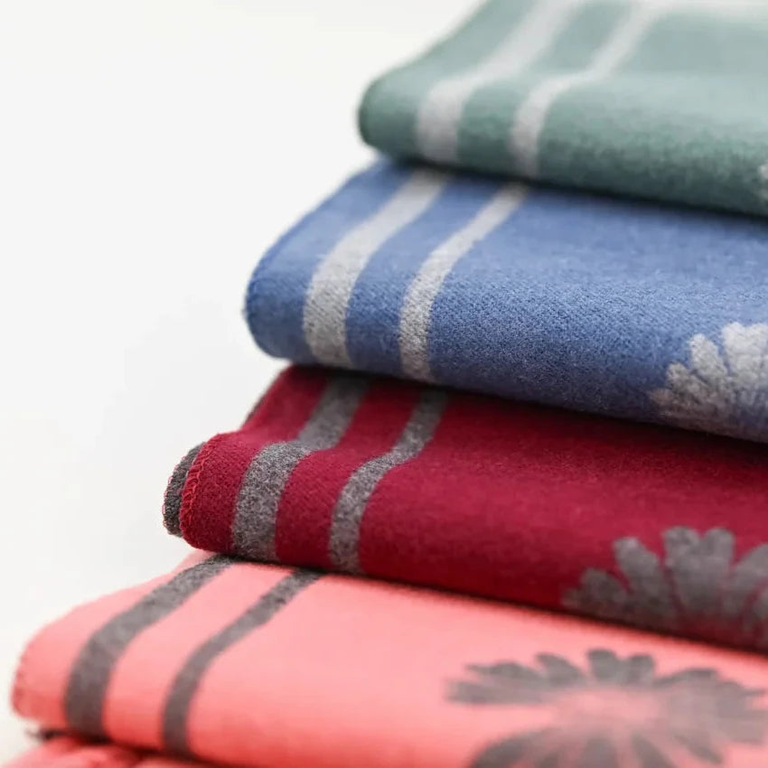 Four folded fabric items stacked diagonally, showing different colors and stripe patterns: sage green, medium blue, deep red, and coral pink, each with a subtle floral motif.